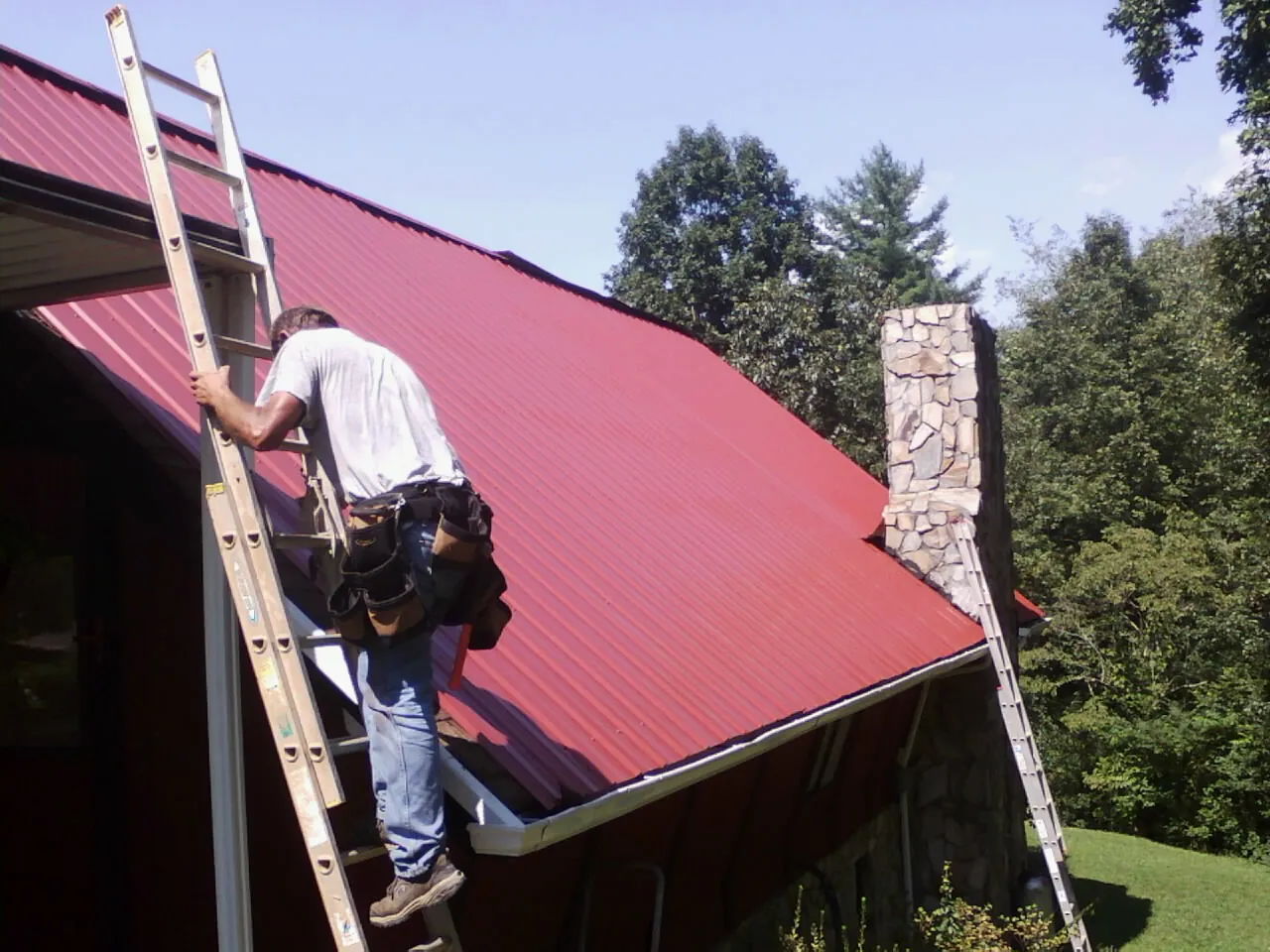 Roofer on a ladder beside a red metal roof and stone chimney — Soffit & Fascia Repair by Roofing Brothers in Inver Grove Heights